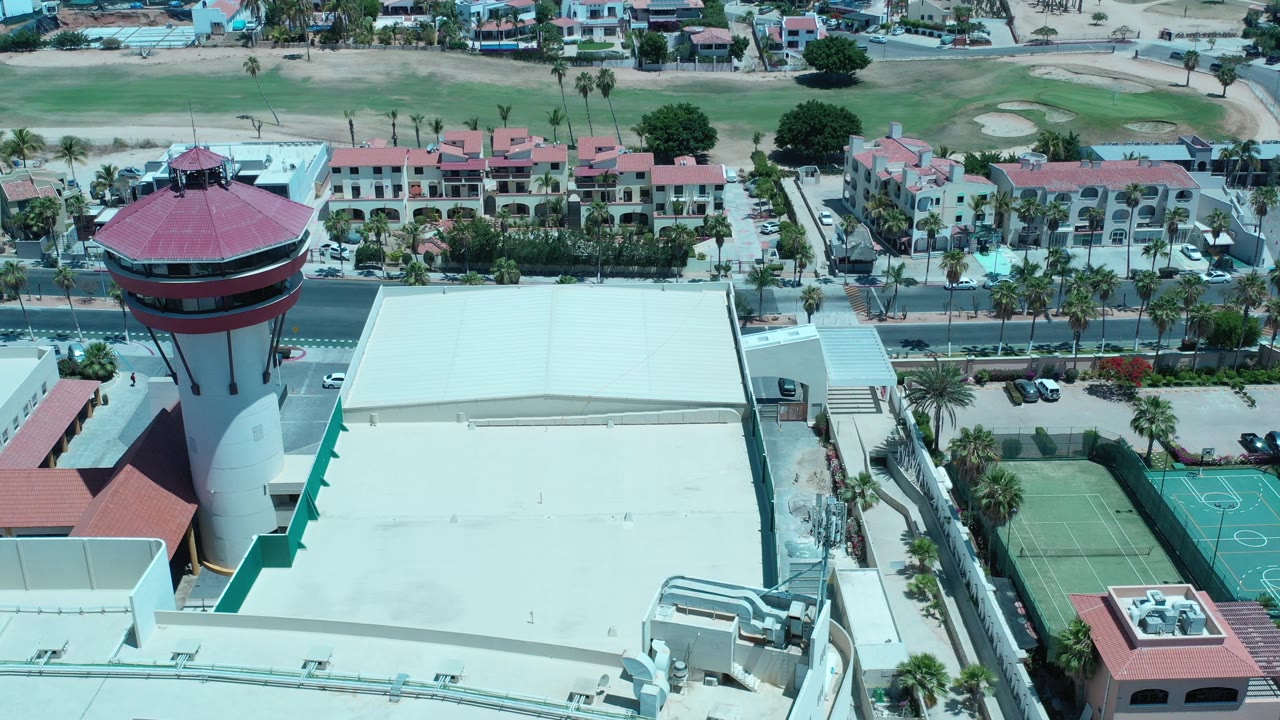 Aerial view of a red circular tower, large pale roof, tennis courts, road, and red-roofed buildings.