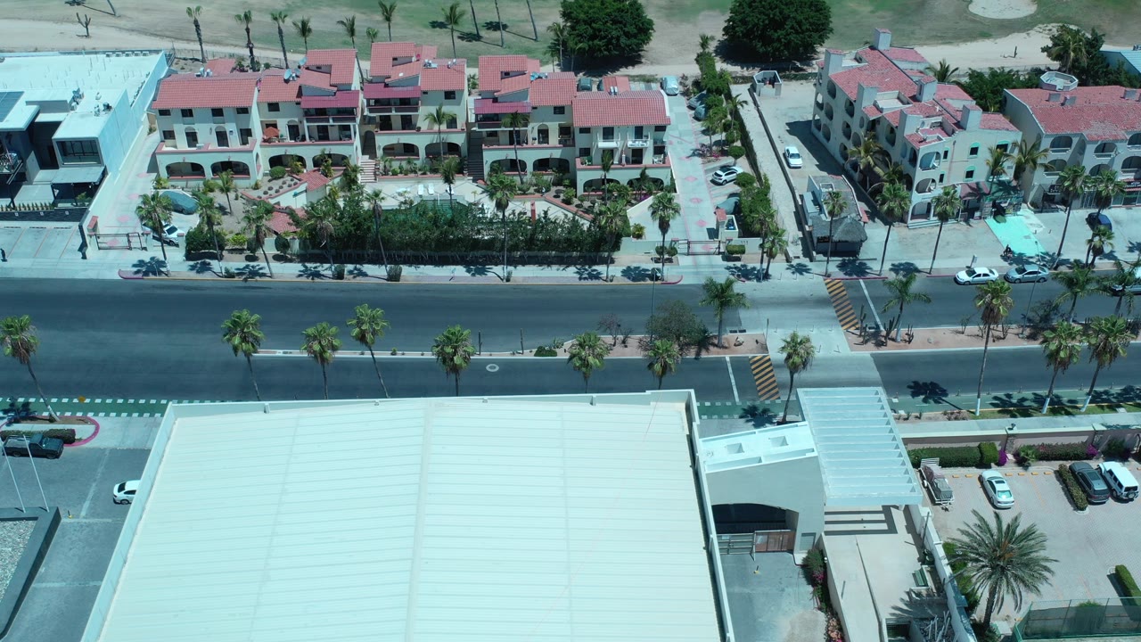 Aerial view in Los Cabos, Mexico showing red-roofed residential buildings above a wide road with palms, parked cars, sidewalks, and no clearly visible people.
