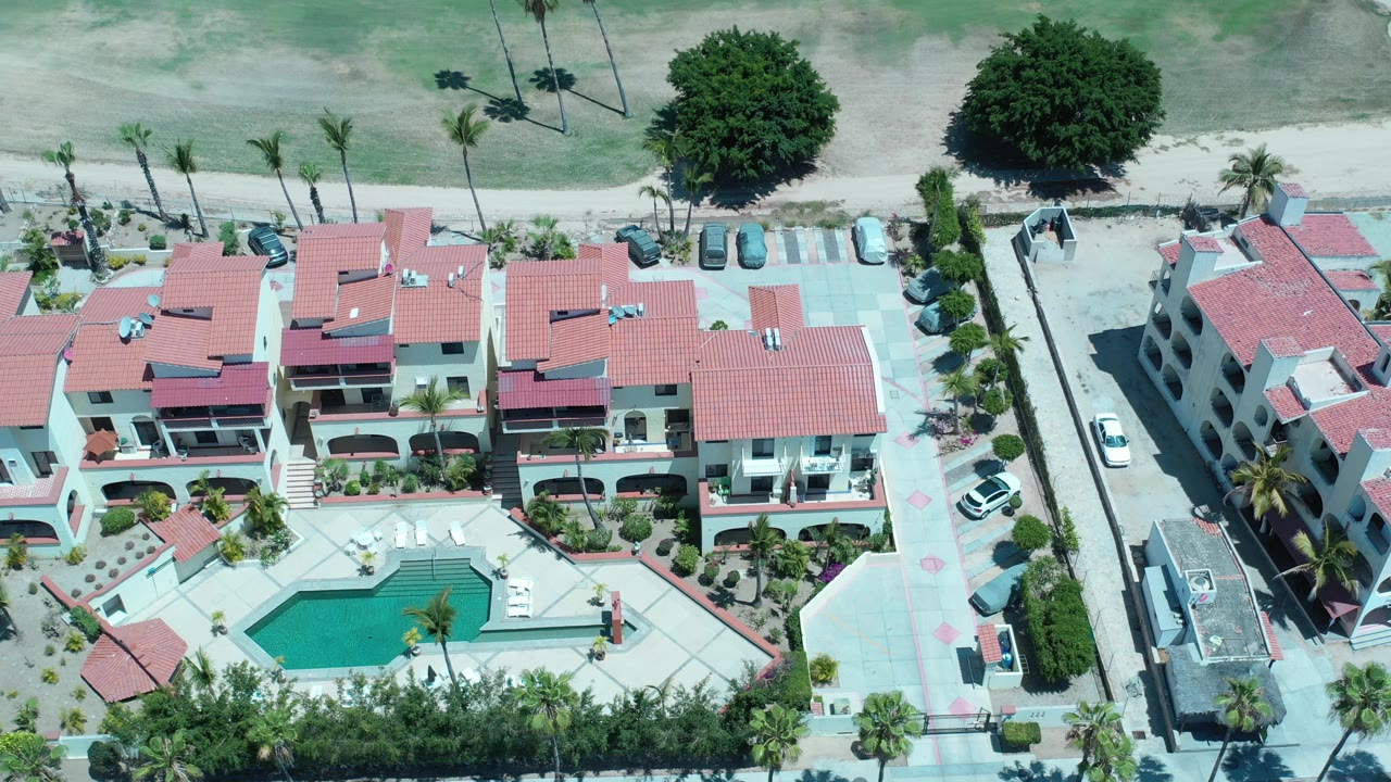 Opening overhead view in Los Cabos, Mexico, showing red tile residential buildings, a turquoise pool, palms, paved courtyard areas, nearby cars, and no visible people.