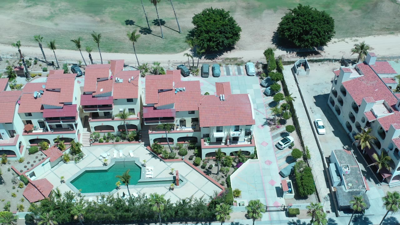 Elevated Los Cabos frame with the pool courtyard lower in view, red roof homes across the center, palms and parked cars along the edges, and no visible people.