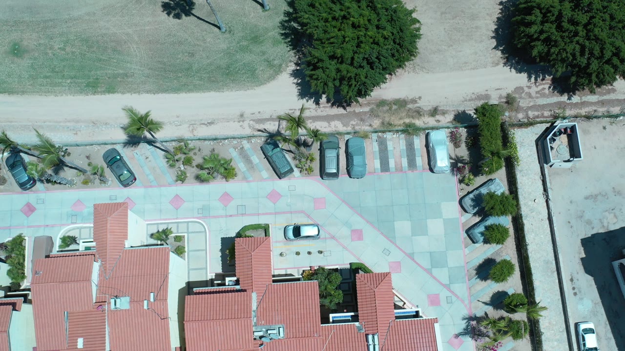 Top-down aerial frame in Los Cabos, Mexico showing red roof apartment buildings, a pale patterned parking area, parked cars, palm trees, a dirt road, and no visible people.