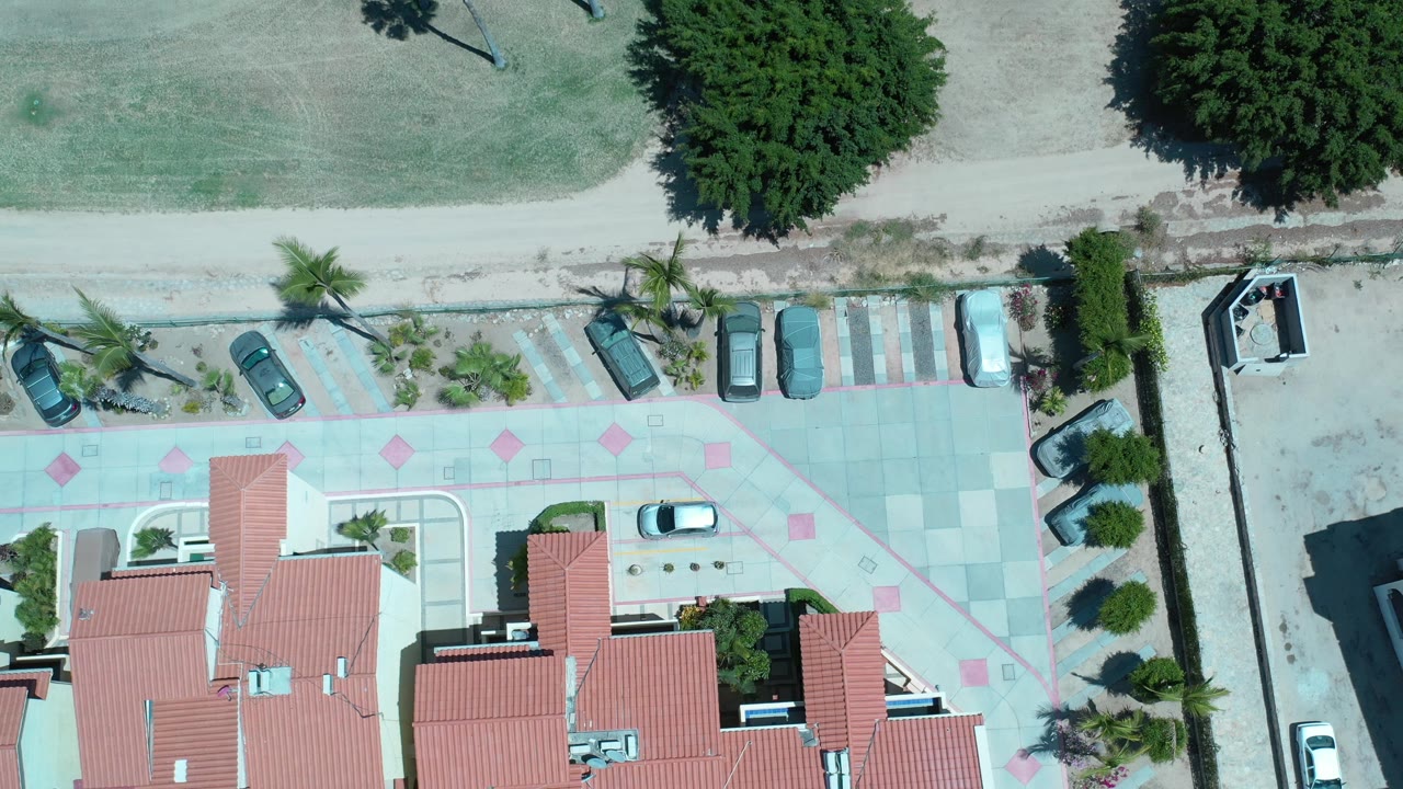 Overhead Los Cabos frame with the apartment roofs centered lower in view, parked cars along the paved lot, a dirt road above, dry grass beyond it, and no visible people.