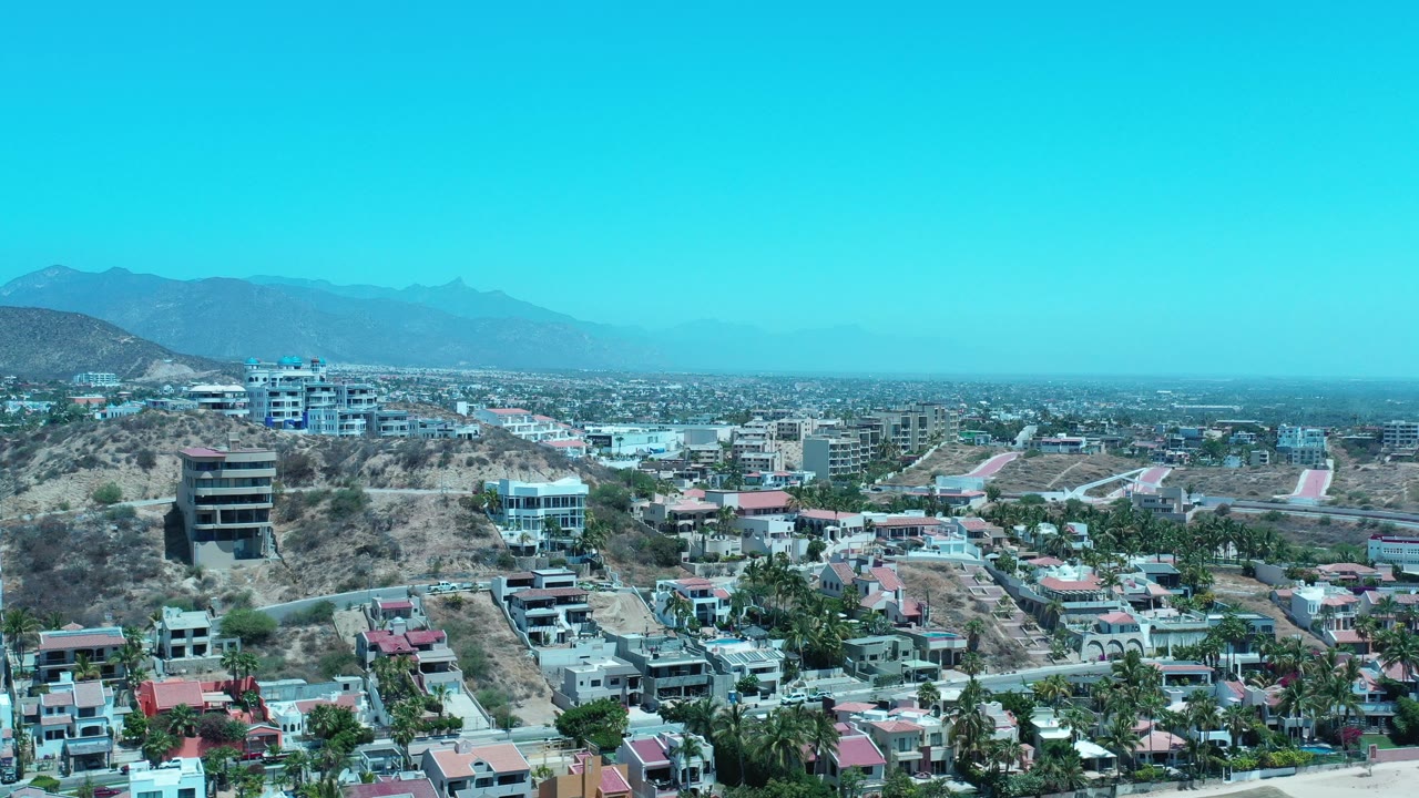 Opening aerial frame over Los Cabos, Mexico, showing rocky hillside homes in the foreground, town buildings across the valley, hazy mountains beyond, and no visible people.