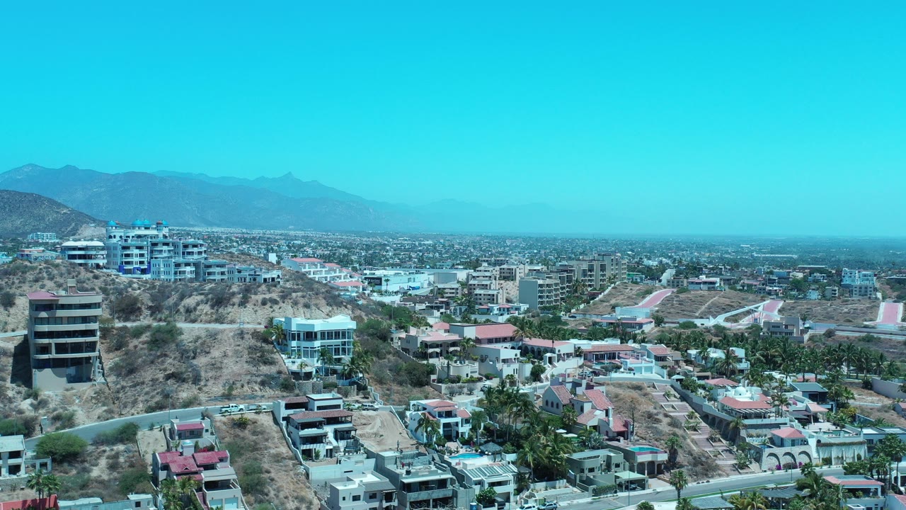 Opening aerial frame over Los Cabos, Mexico with hillside residences in the foreground, dense city development beyond, hazy mountains on the horizon, bright blue sky, and no visible people.