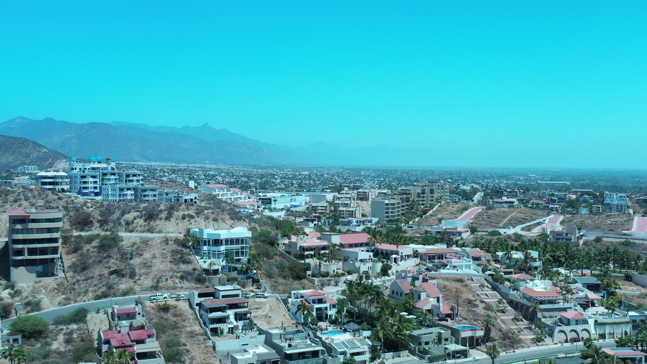 Elevated view of hillside residences, a road, city blocks, mountains, and blue sky.