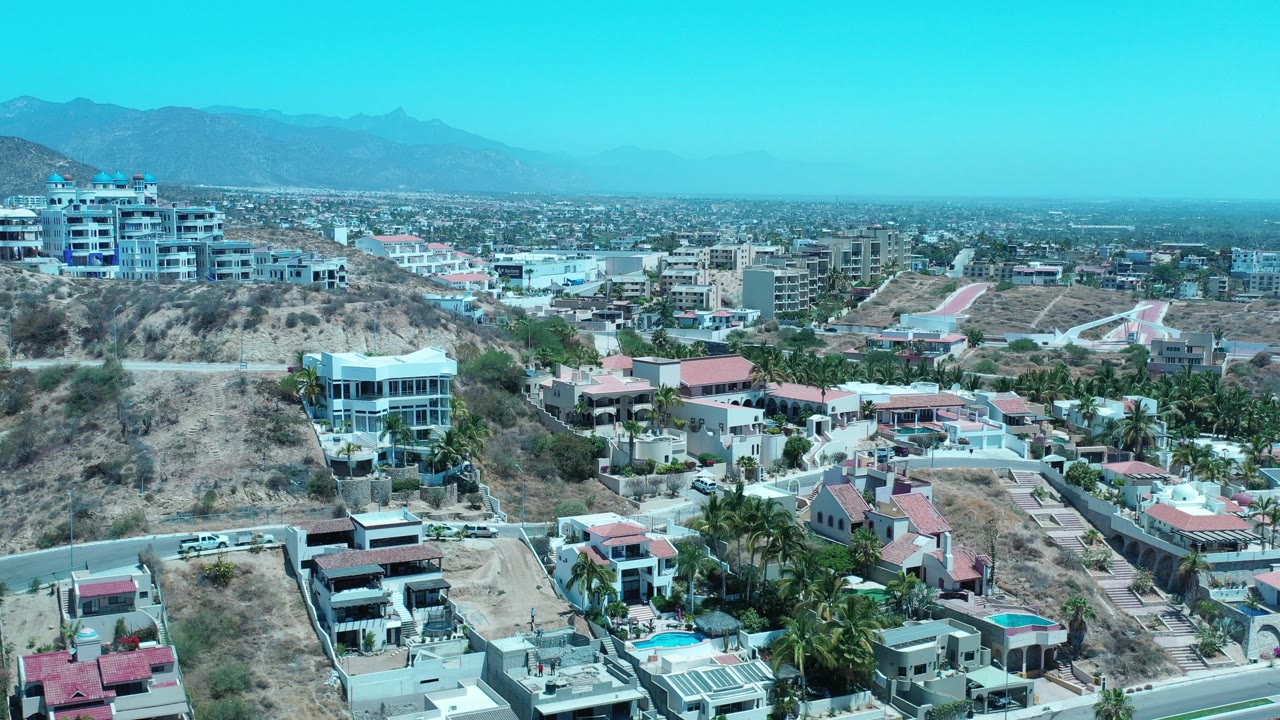Opening aerial frame of Los Cabos, Mexico with modern homes below, larger hillside buildings at left, a broad city area beyond, hazy mountains, and no visible people.