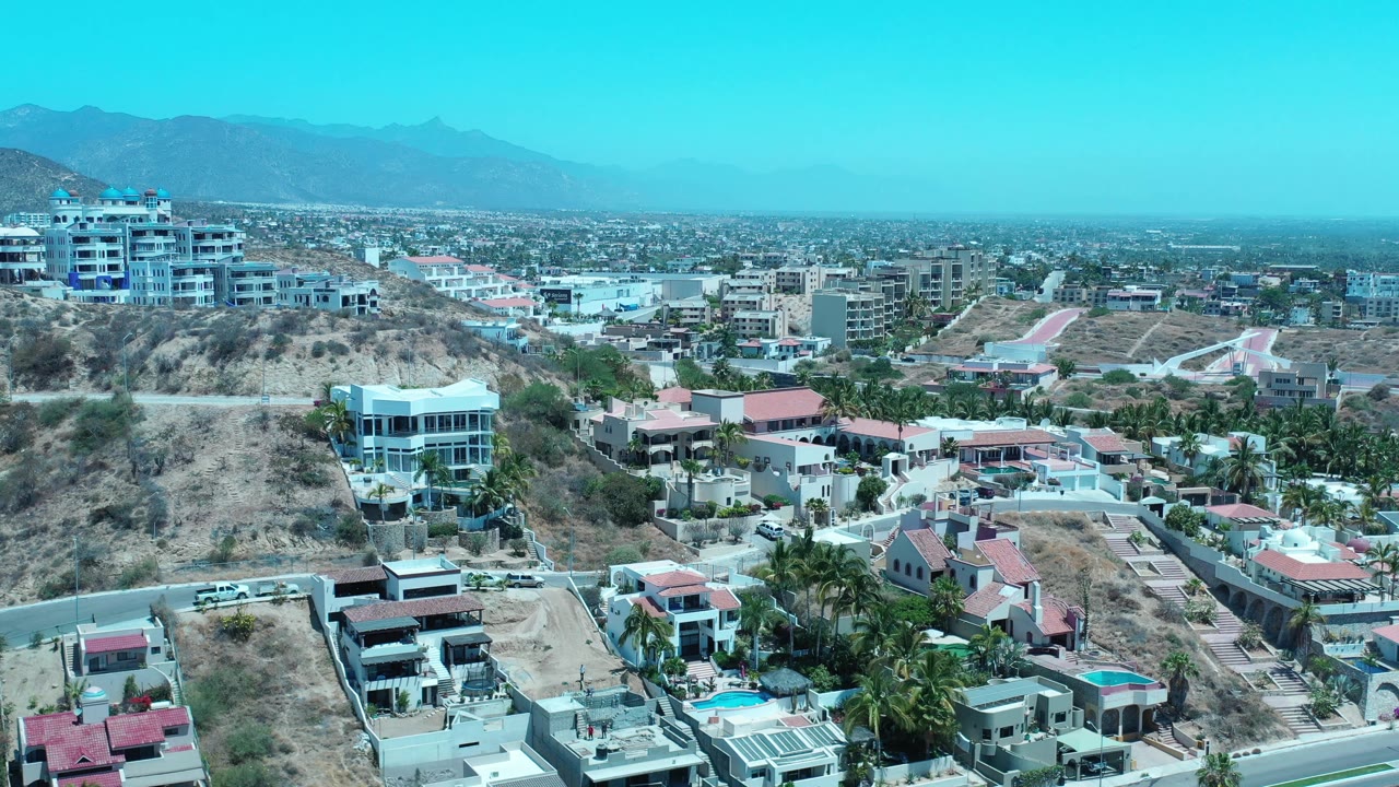 Aerial frame of Los Cabos, Mexico with a white multi-story building on the left hillside, red-roofed homes below, roads in the foreground, distant buildings, and no visible people.
