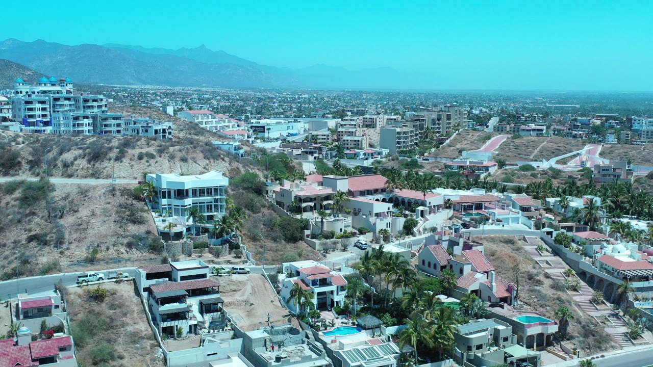 Aerial view across Los Cabos homes, hillside buildings, and a hazy mountain horizon.