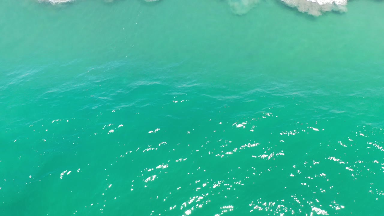 Aerial view of turquoise ocean water in Los Cabos, Mexico, with white breaking surf near the top edge and no visible people.