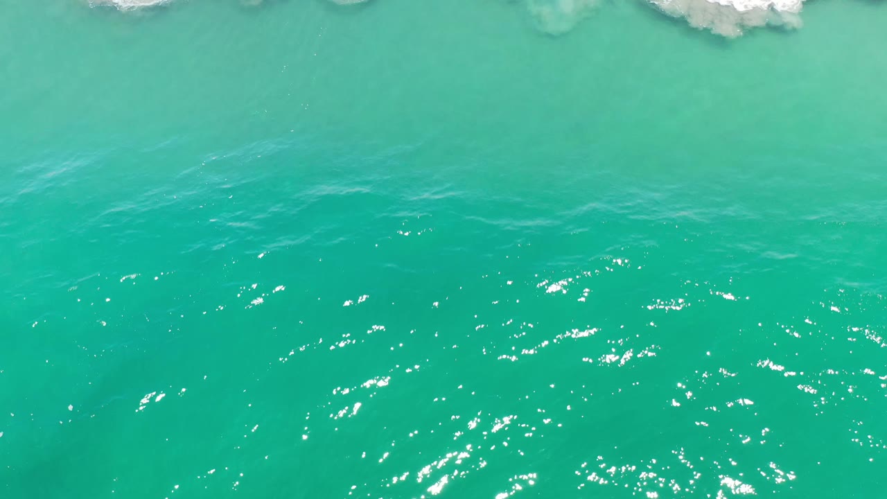 Aerial Los Cabos ocean frame with sunlit blue-green water, scattered surface sparkle, and a narrow band of wave foam, no visible people.