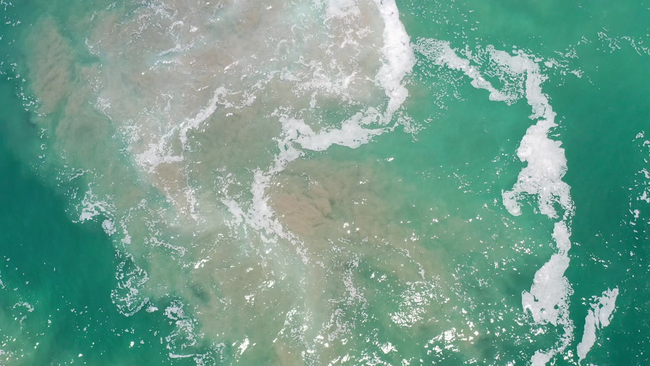 Overhead view of green ocean water and white foam over pale sand in Los Cabos, Mexico, with no visible people.