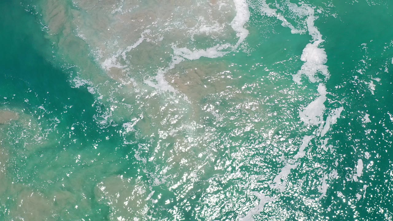 Overhead view of emerald water in Los Cabos, Mexico, with white foam bands, bright reflected sunlight, tan shallows, and no visible people.