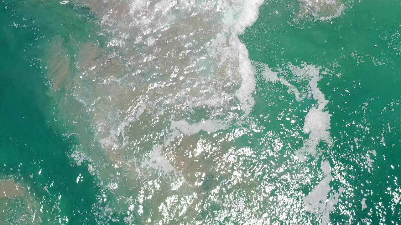 Aerial view of Los Cabos ocean water with scattered white bubbles, glinting light on the surface, muted sand visible below, and no visible people.