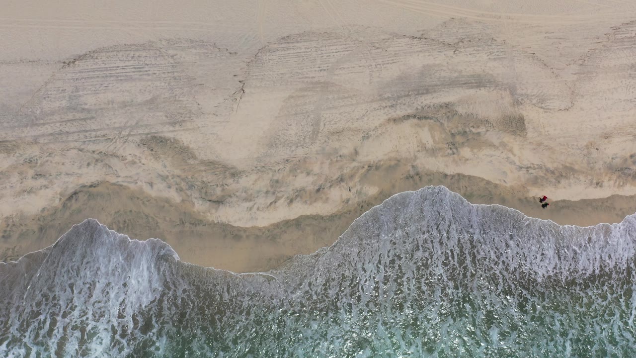 Top-down view of white surf reaching pale sand in Los Cabos, Mexico, with wet shoreline patterns and no visible people.