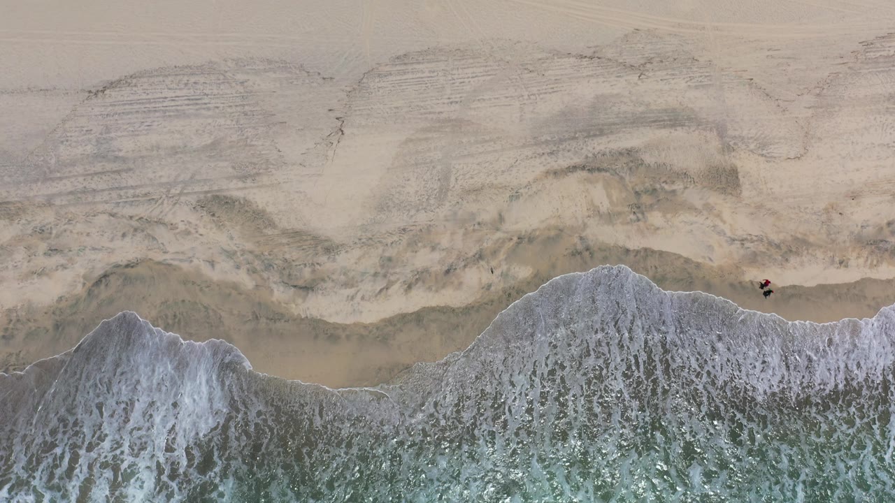 Aerial frame of foamy water spread across the Los Cabos shoreline, with rippled wet sand above and no visible people.