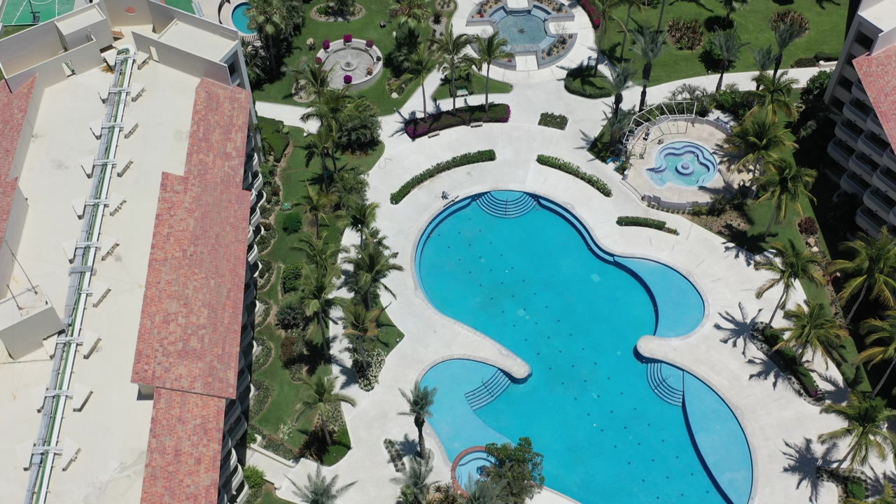 Aerial view in Los Cabos, Mexico showing a large turquoise pool, pale deck, palms, lawns, and a hotel building along the left edge with no visible people.