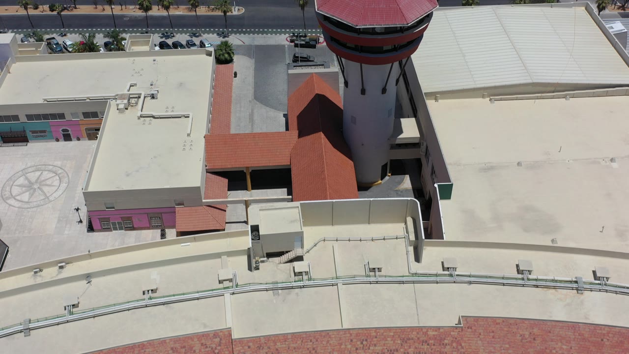 Top-down Los Cabos, Mexico frame with cream concrete rooftops, a red octagonal tower roof, a road at the top edge, parked cars, palms, and no visible people.