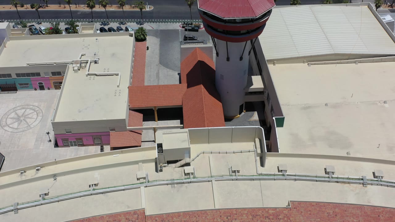 Los Cabos overhead view showing the red-roofed tower shifted toward the right, cream roof surfaces, a small parking area, roadside palms, cars, and no visible people.