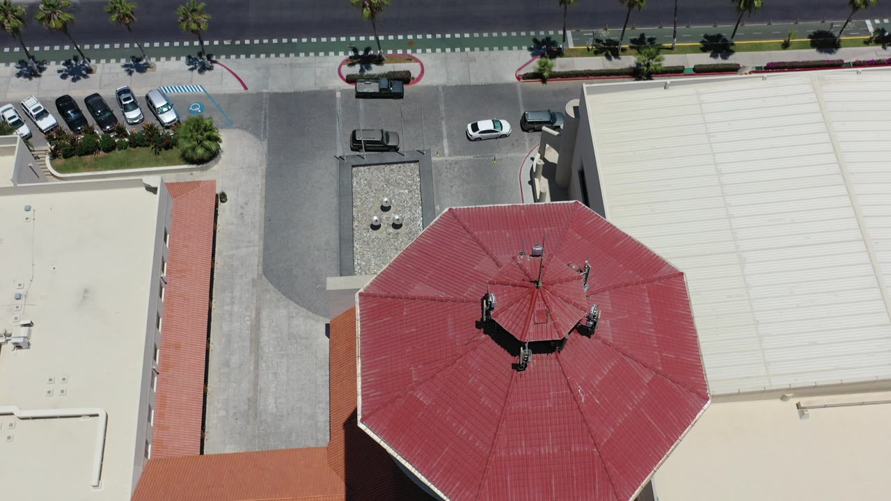 Top-down aerial frame in Los Cabos, Mexico showing a red octagonal roof beside light building roofs, a parking lot, a road with cars, palm plantings, and no visible people.