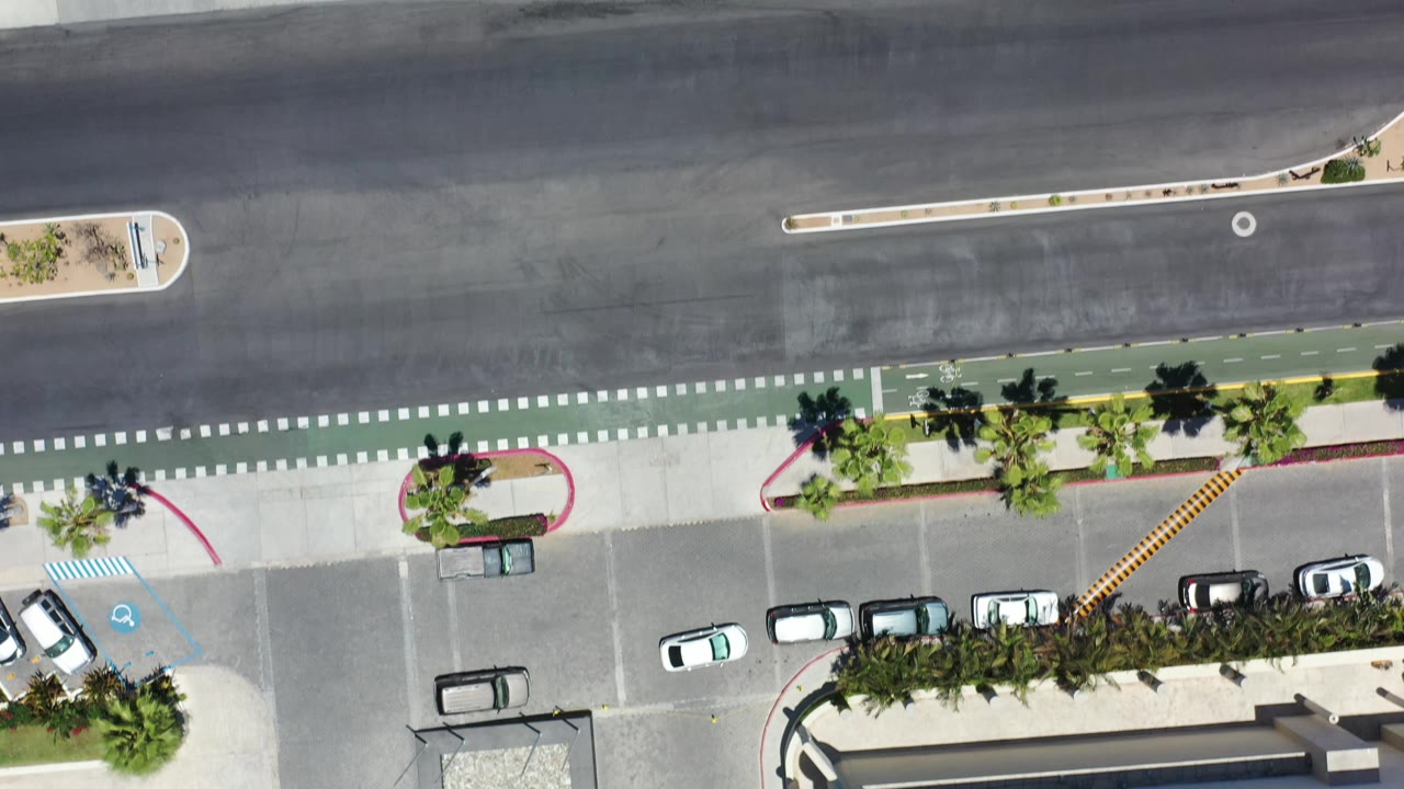 Overhead Los Cabos resort approach with a broad asphalt road, green bike lane, curb islands, sidewalk, parking bays, palms, and no visible people.