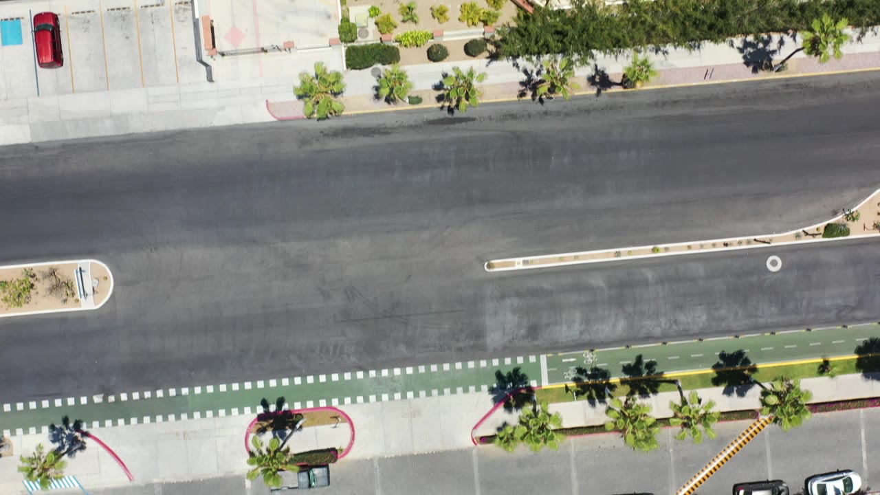 Vertical aerial frame in Los Cabos, Mexico showing resort frontage beside a wide road, a green bike lane, landscaped palms, sidewalk paths, and no visible people.