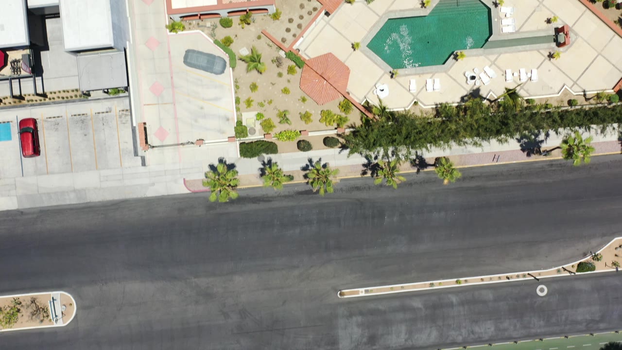 Top-down Los Cabos resort view with a turquoise pool, red tile roofs, paved road, parking spaces, desert landscaping, shade trees, and no visible people.