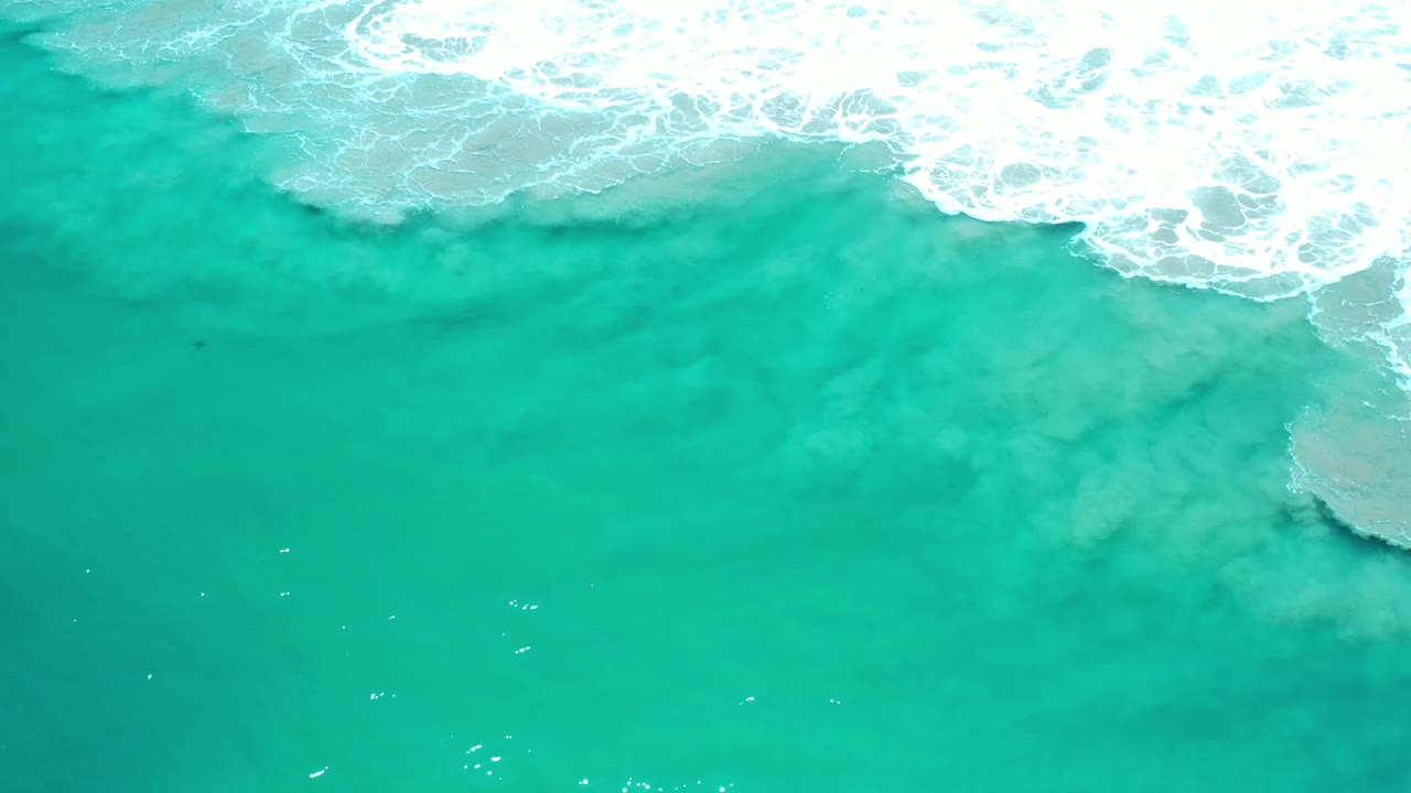 Overhead view of turquoise-green seawater in Los Cabos, Mexico, with white surf foam running across the upper edge and no visible people.