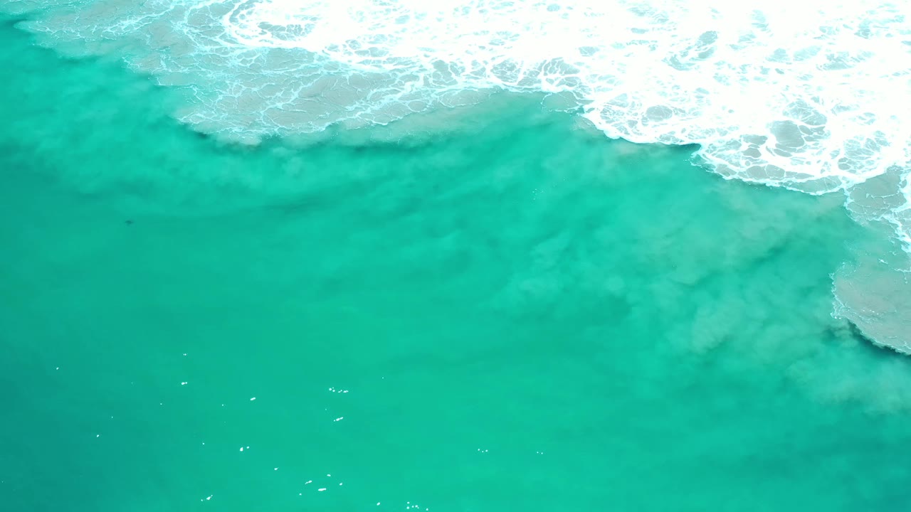 Overhead Los Cabos ocean frame with white wave bubbles spreading across green water and open empty sea below, with no visible people.