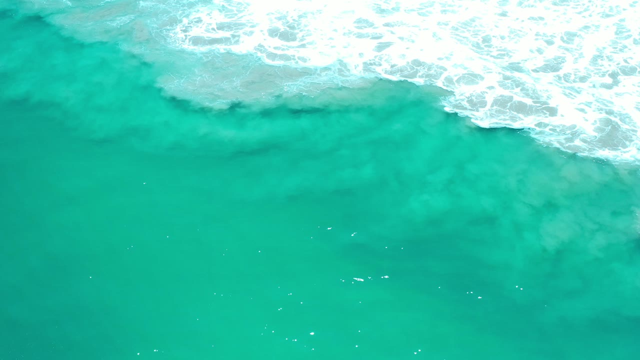 Overhead view of turquoise-green water in Los Cabos, Mexico, with a wide band of white surf across the top and no visible people.