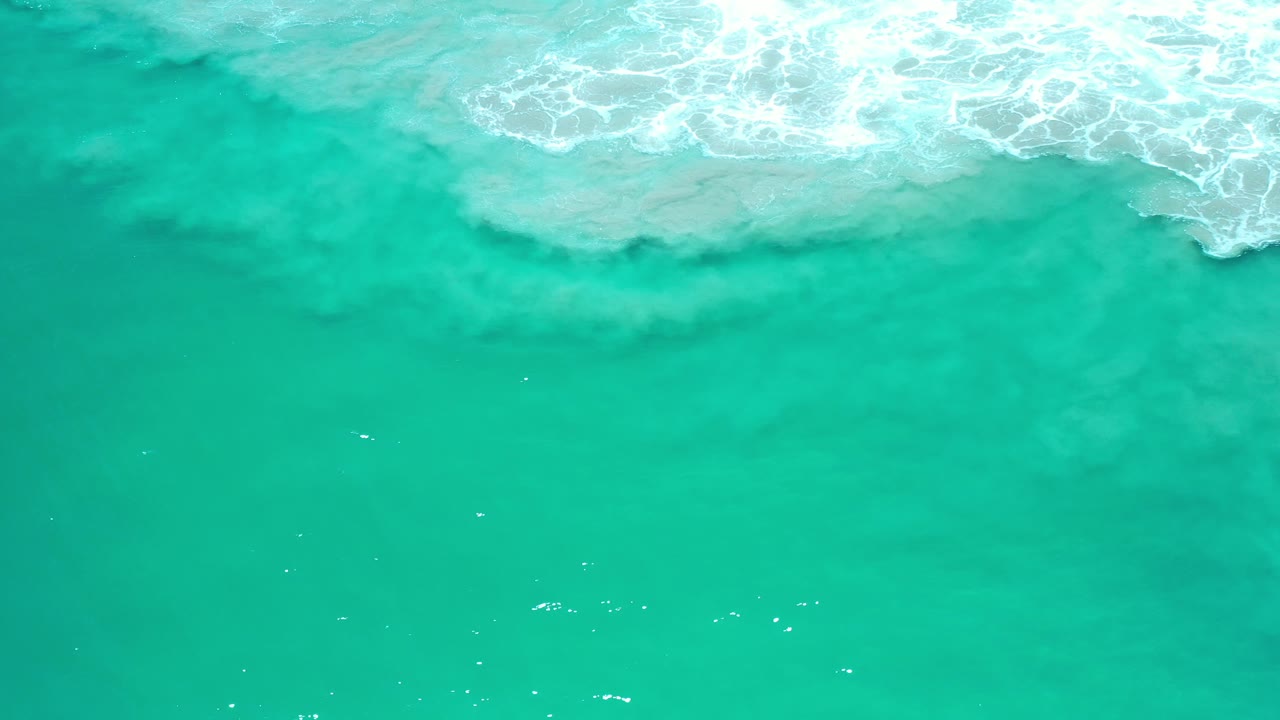 Overhead view of turquoise ocean water in Los Cabos, Mexico, with white surf foam entering along the upper edge and no visible people.