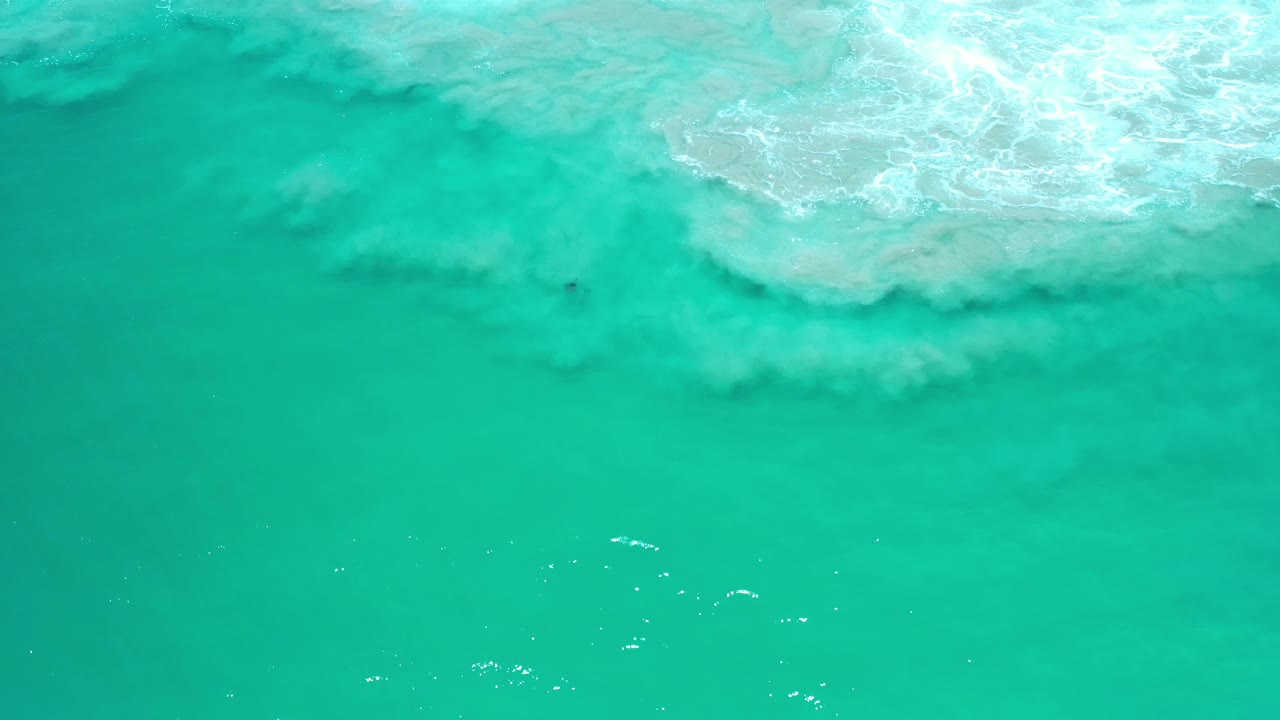 Gray-white surf churns across the upper right over bright teal ocean.