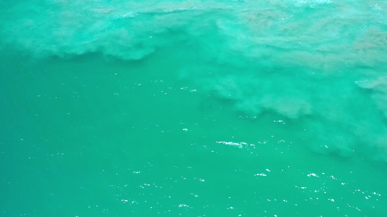 Opening frame of clear green water over pale loose sand in Los Cabos, Mexico, with small sun glints and no visible people.