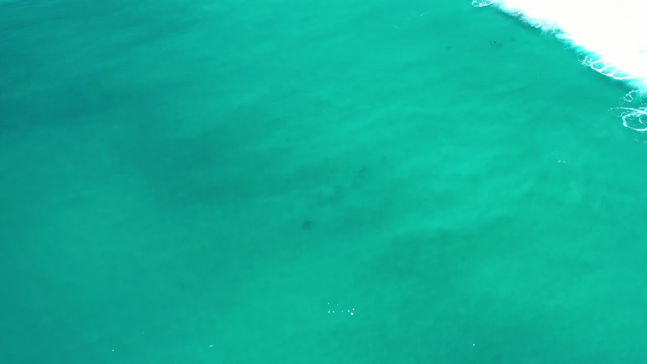 Overhead view of teal sea in Los Cabos, Mexico, with a band of white foam entering from the upper-right corner and no visible people.