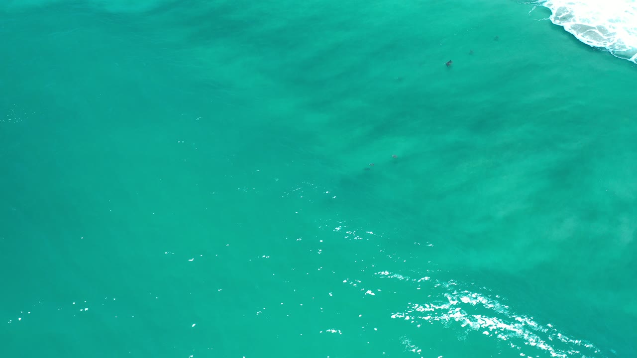 Overhead view of turquoise ocean water in Los Cabos, Mexico, with faint darker patches below the surface, sun sparkle near the lower center, white surf at the upper right edge, and no visible people.
