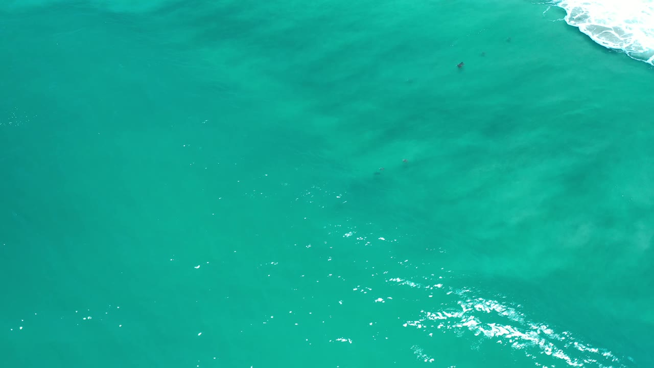 Aerial Los Cabos ocean frame with open teal water across most of the image, a thin white surf line along the upper right, subtle darker shapes under the water, and no visible people.