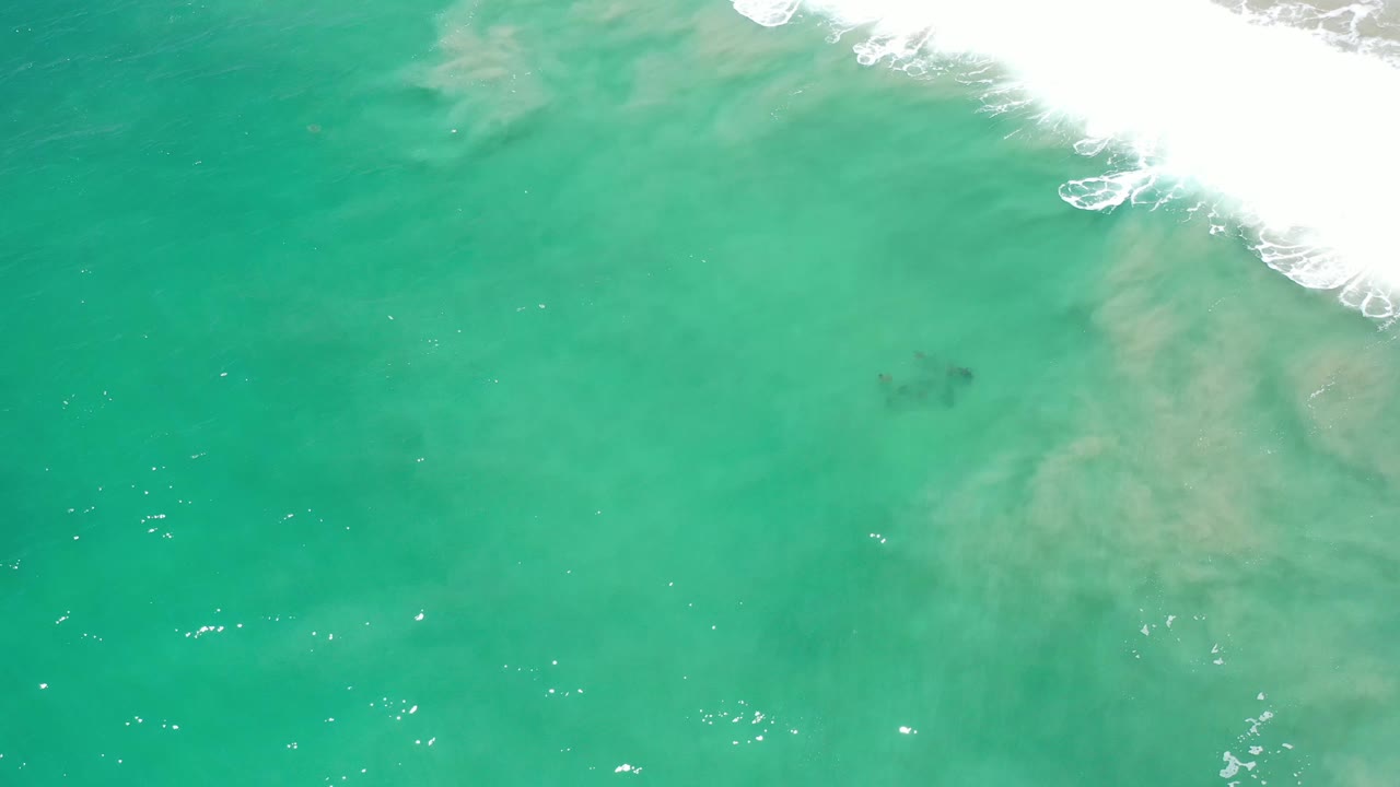 Aerial-looking frame over Los Cabos water, with a bright foam line near the top right and faint tan sand shapes under the surface, no visible people.