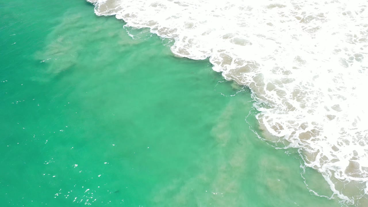 Overhead view in Los Cabos, Mexico of turquoise-green water with white surf foam pushing in from the upper right, with no visible people.