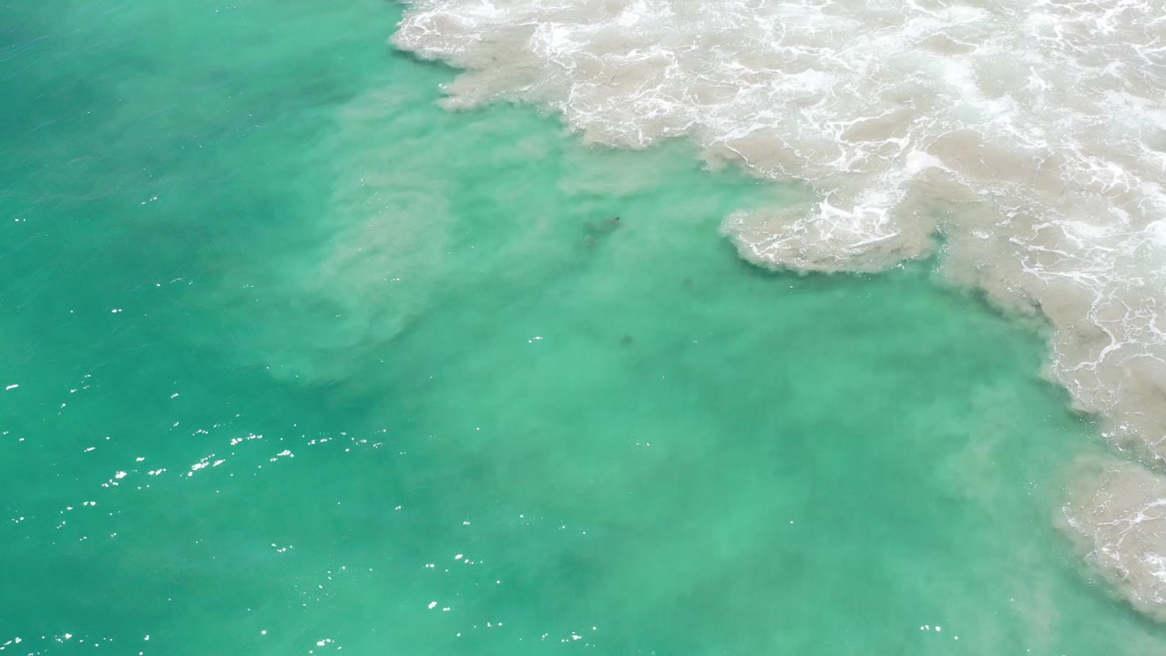 Turquoise water beside white surf on pale sand.