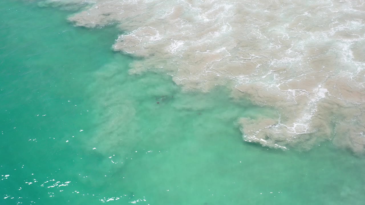 Overhead view in Los Cabos, Mexico, with turquoise water at left and pale foamy shallows along the upper right, no visible people.