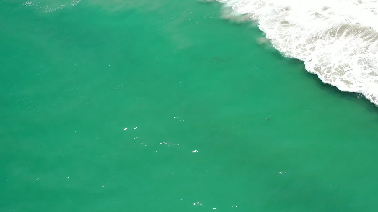 Aerial view of turquoise-green ocean in Los Cabos, Mexico, with white surf beginning to enter from the upper right and no visible people.