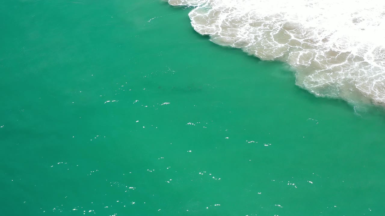 Green ocean surface in Los Cabos, Mexico, with bright white foam spread across the upper right and scattered highlights on the water, no visible people.
