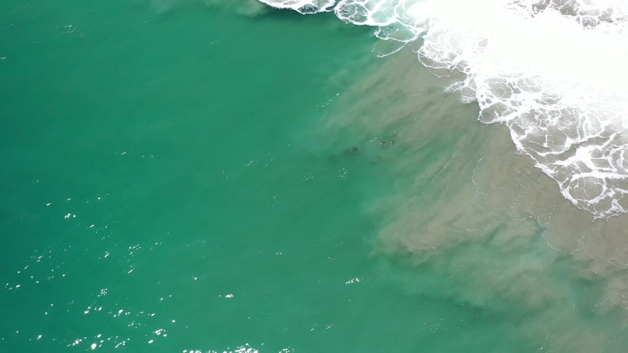 Overhead view in Los Cabos, Mexico, showing green seawater, tan sand beneath the surface, white foam at the upper right, and no visible people.