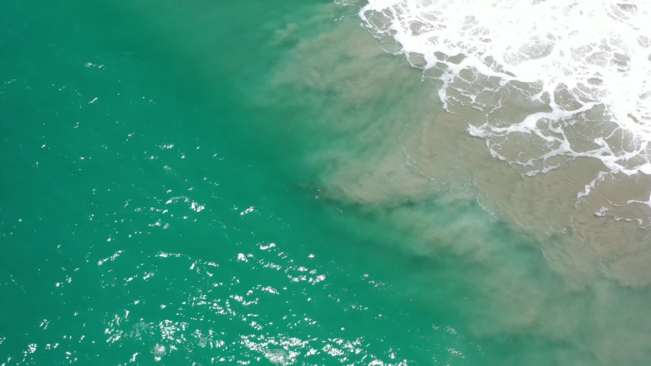 Aerial view over green-blue ocean in Los Cabos, Mexico, with white surf foam at the upper right, sandy water beneath the wash, sun glints on the surface, and no visible people.