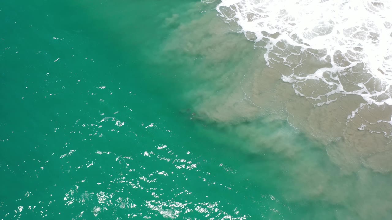 Top-down Los Cabos ocean frame with emerald water, scattered sunlight reflections, pale sand visible through the shallows, white foam along the upper right, and no visible people.