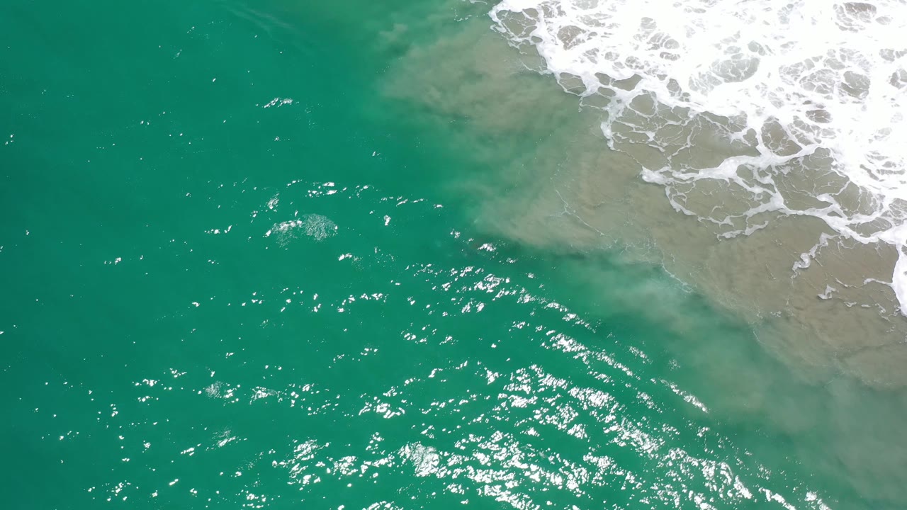 Overhead view of green water in Los Cabos, Mexico, with stronger sun sparkle across the center, pale sandy turbulence at the upper right, white breaking foam, and no visible people.