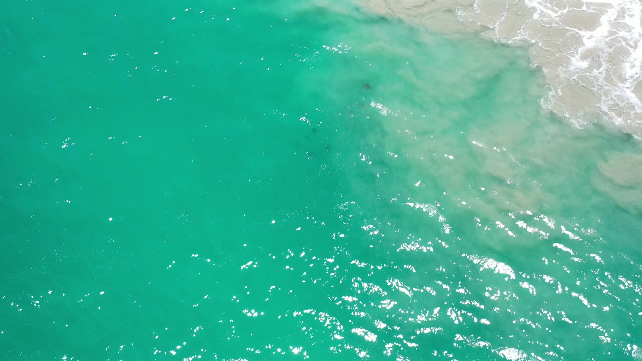 Overhead view of turquoise-green water in Los Cabos, Mexico, with white surf and pale sandy wash at the upper right, bright sun glints on the ripples, and no visible people.