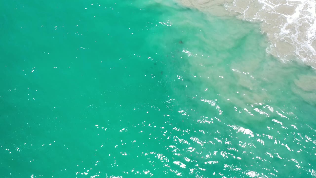 Aerial frame over Los Cabos water where green-blue surface ripples fill most of the view and foamy shoreline wash sits near the upper right edge, with no visible people.