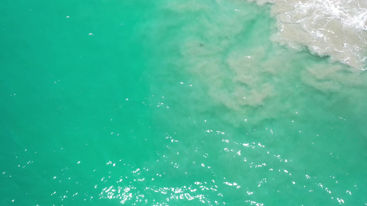 Overhead Los Cabos water view with turquoise color, scattered sun sparkle, and foamy surf holding near the upper right, with no visible people.