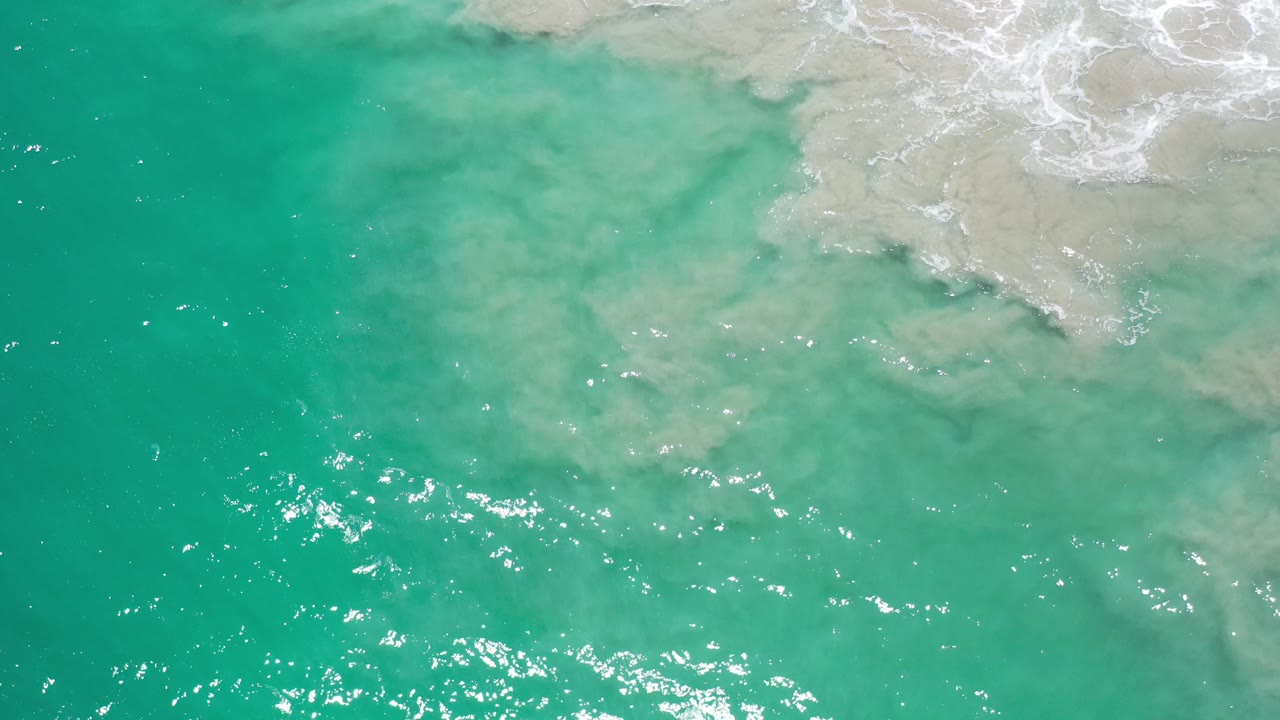 Overhead view in Los Cabos, Mexico, with turquoise water across the lower frame, pale sand and foam at the upper right, and no visible people.
