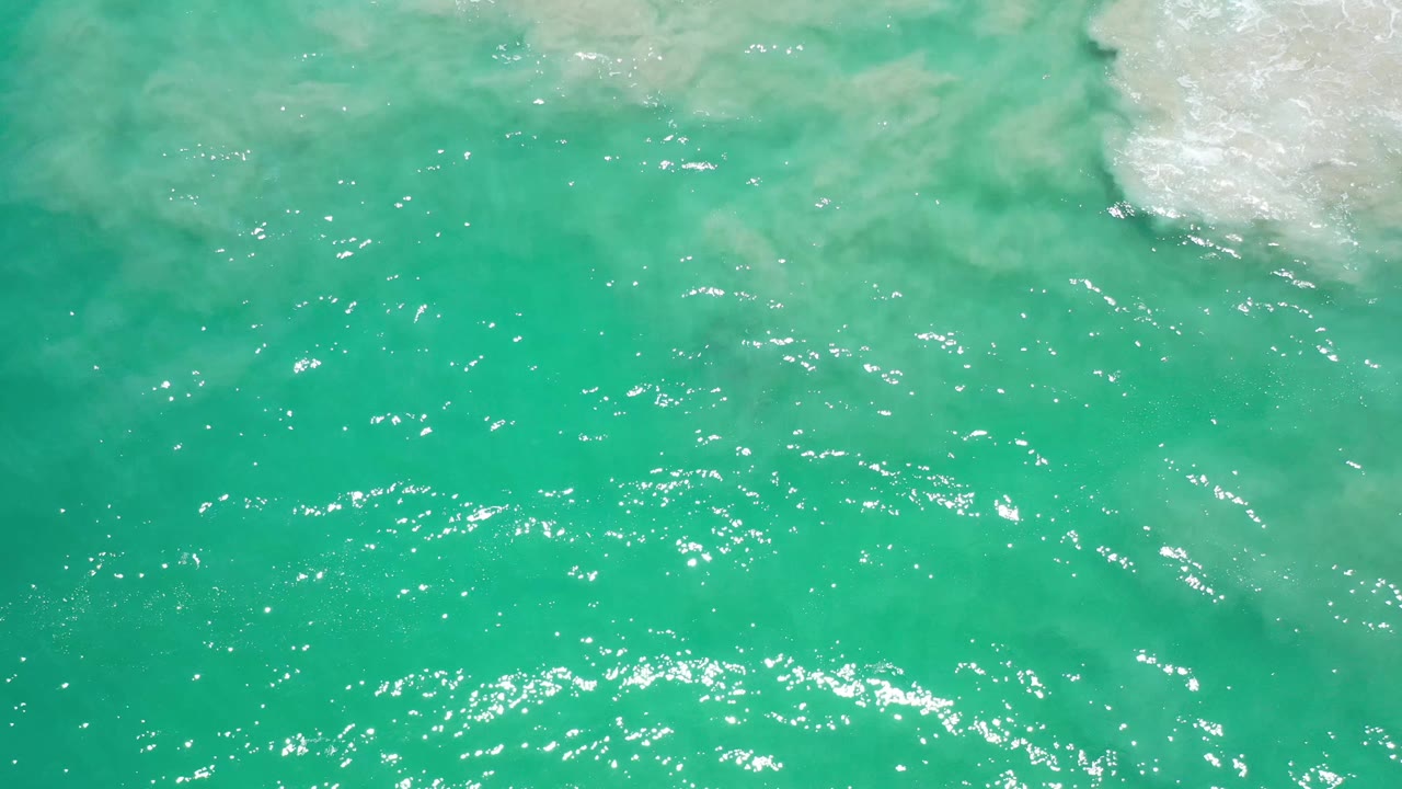 Overhead Los Cabos ocean frame with green water, scattered white sparkle across the ripples, a pale foam patch near the upper right, and no visible people.